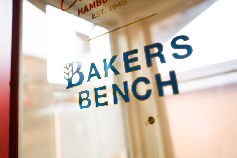 An artful image of the glass door leading into Bakers Bench; a sign on the door reads “Bakers Bench” in a sans-serif typeface